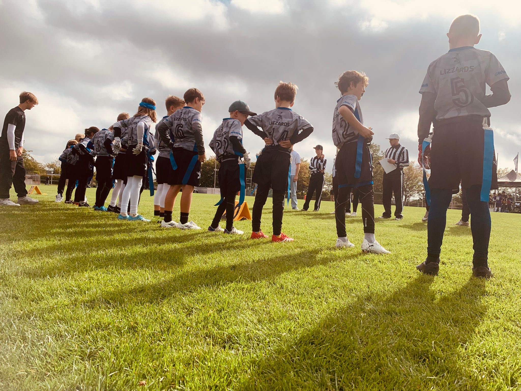 Ein Foto aus niedriger Perspektive zeigt die U10-Mannschaft der Kelkheim Lizzards, die in einer geraden Linie mit dem Rücken zur Kamera steht und den Schiedsrichtern auf der anderen Seite des Spielfelds zugewandt ist.
