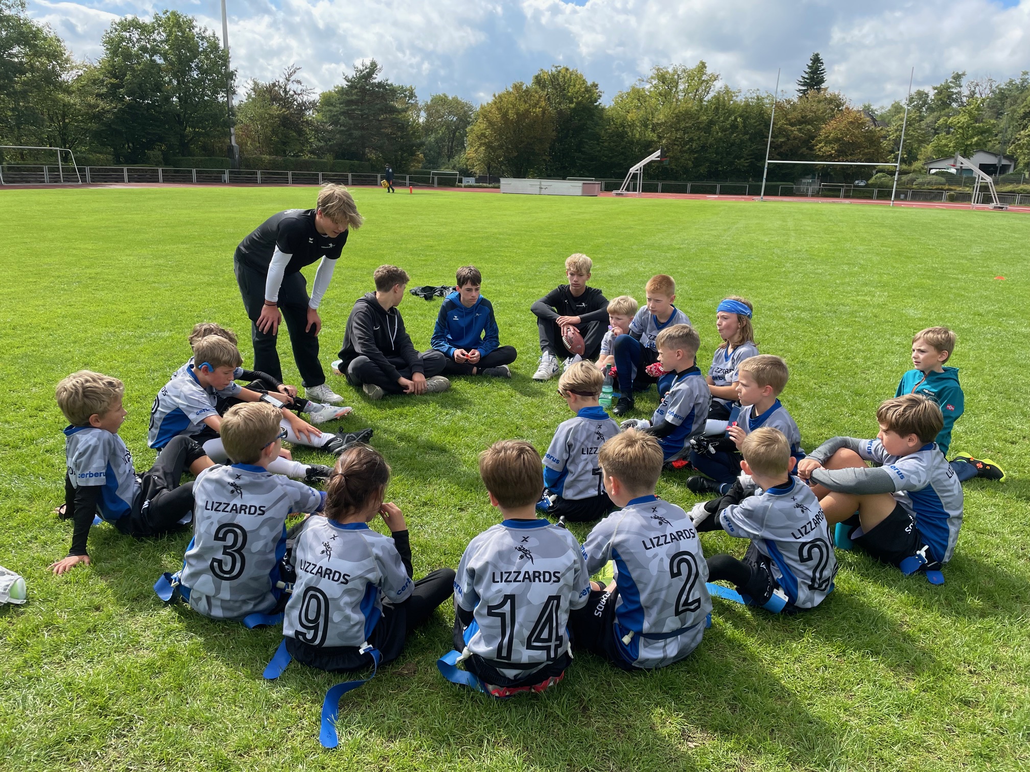 Die Kelkheimer Lizzards U10 sitzen im Kreis auf dem Feld und hören ihrem Trainer zu.