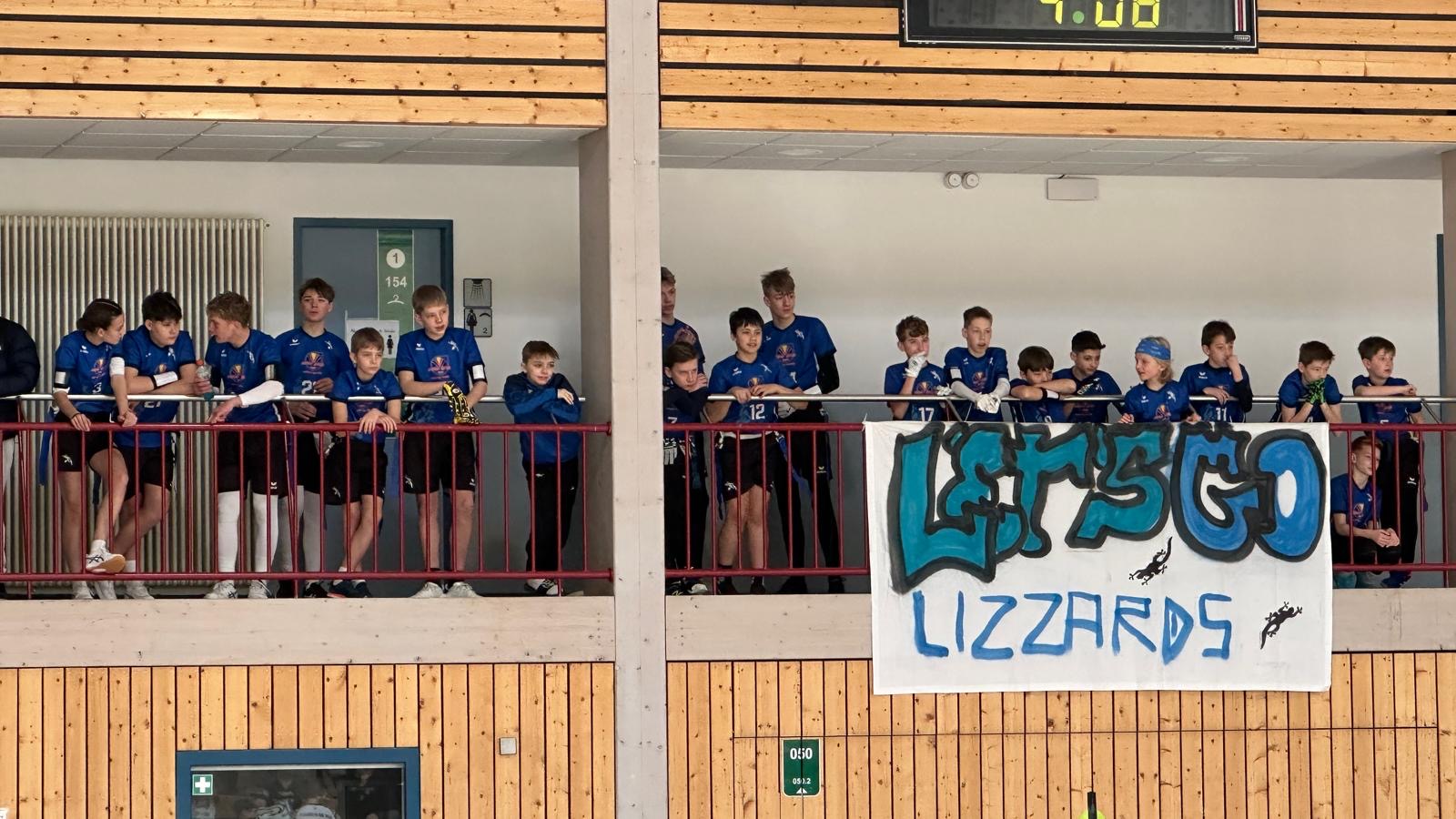 Lizzards-Spieler verfolgen ein Spiel von der Tribüne aus neben einem Banner mit der Aufschrift „Let's Go Lizzards”.