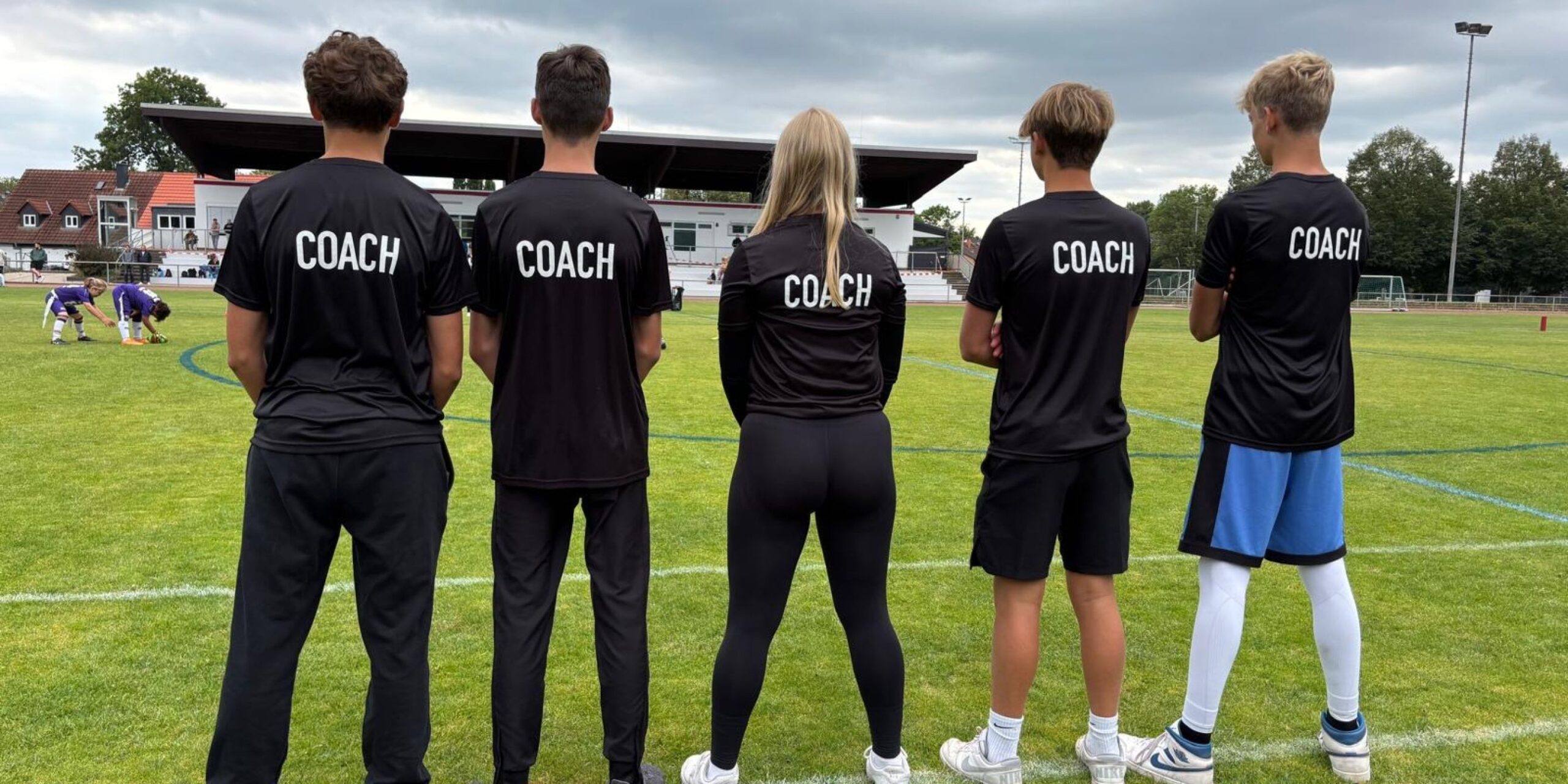 Fünf Trainer stehen mit dem Rücken zur Kamera in einer Reihe und beobachten die Spieler beim Training auf dem Feld.