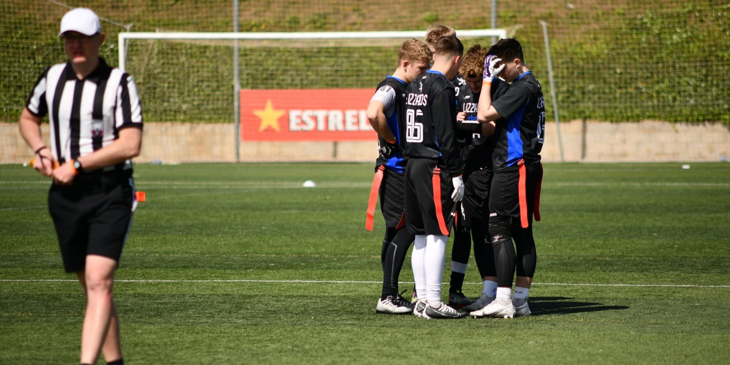 Erfahrene Spieler stehen in einem engen Kreis auf dem Rasenfeld, während der Schiedsrichter sich von ihnen entfernt.