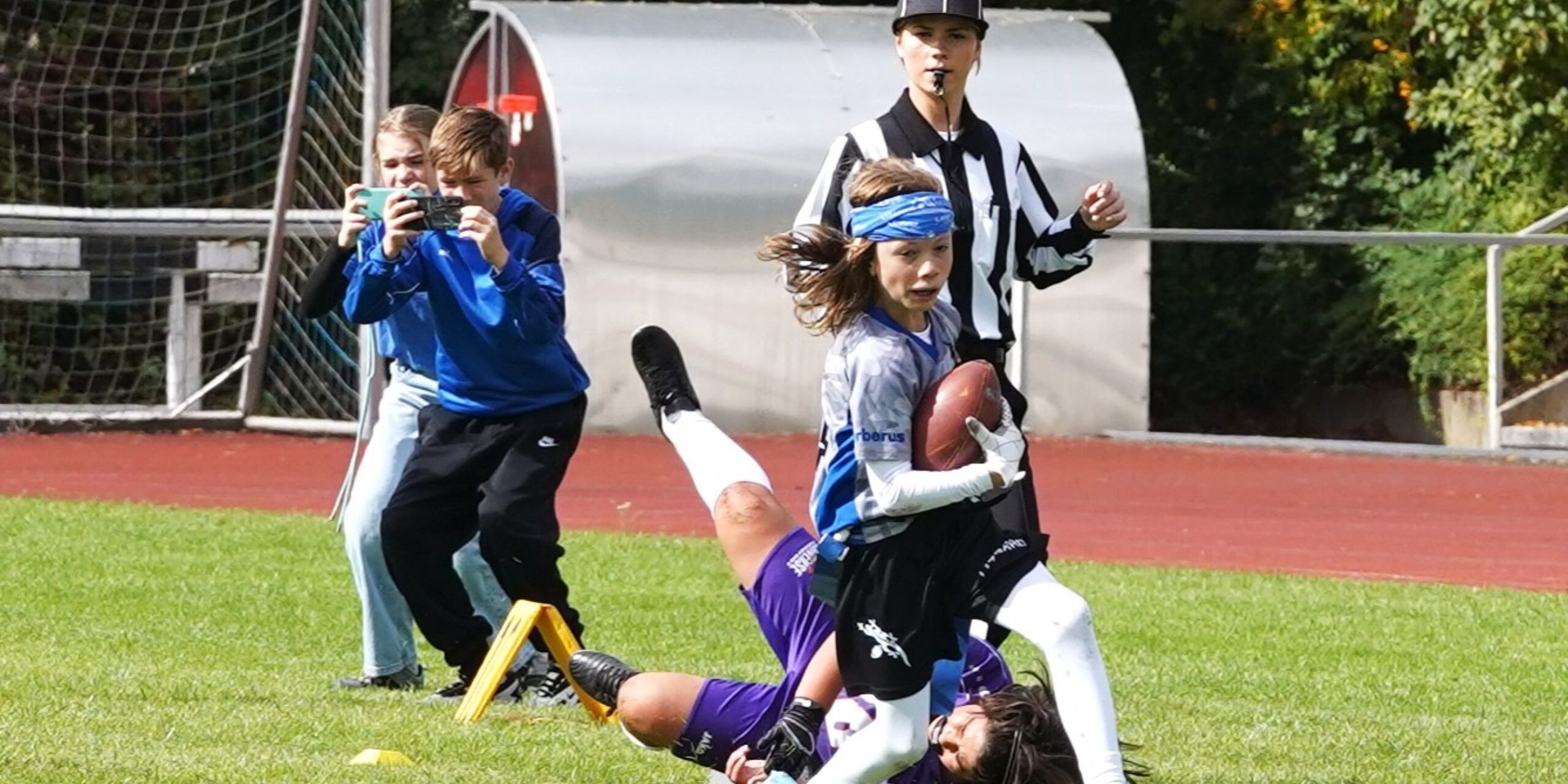 Ein kleiner Junge rennt mit dem Ball, während ein anderer Spieler hinter ihm zurückfällt. In der Nähe steht ein Schiedsrichter, und im Hintergrund machen zwei Teenager Fotos.